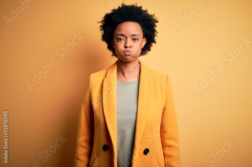 Young beautiful African American afro businesswoman with curly hair wearing yellow jacket puffing cheeks with funny face. Mouth inflated with air, crazy expression. © Krakenimages.com