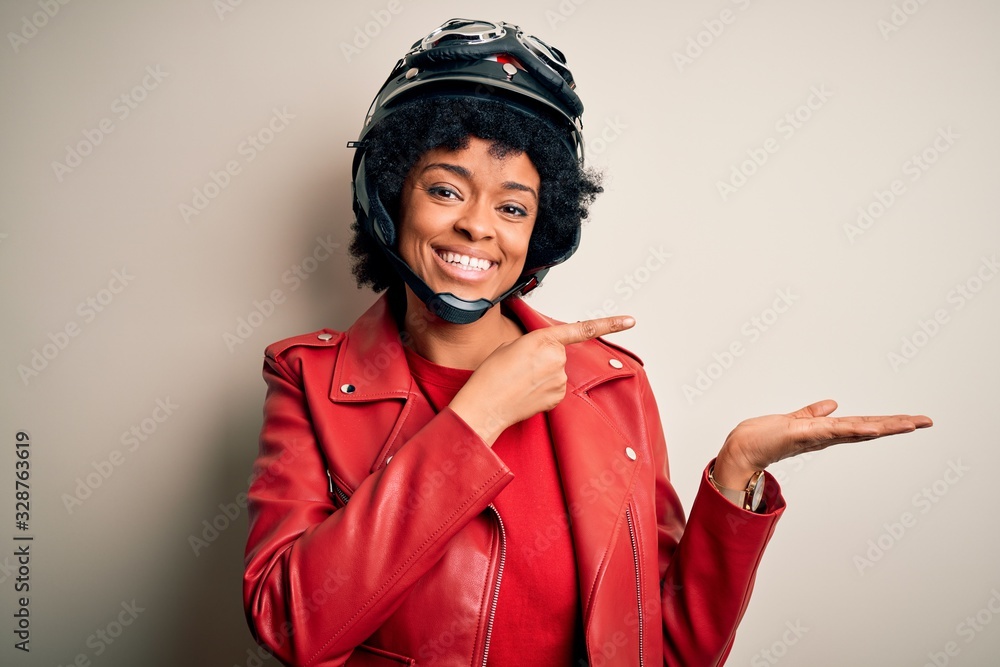 Young African American afro motorcyclist woman with curly hair wearing motorcycle helmet amazed and smiling to the camera while presenting with hand and pointing with finger. Young African American afro motorcyclist woman with curly hair wearing motorcycle helmet amazed and smiling to the camera while presenting with hand and pointing with finger.