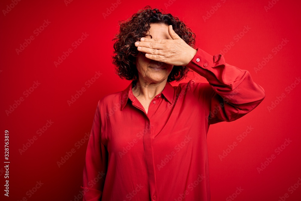 Middle age beautiful curly hair woman wearing casual shirt and glasses over red background covering eyes with hand, looking serious and sad. Sightless, hiding and rejection concept