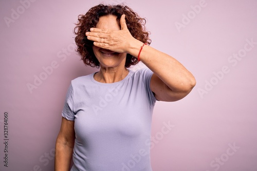 Middle age beautiful curly hair woman wearing casual t-shirt over isolated pink background smiling and laughing with hand on face covering eyes for surprise. Blind concept.