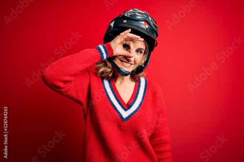 Middle age beautiful blonde motorcyclist woman wearing moto helmet over red background doing ok gesture with hand smiling, eye looking through fingers with happy face.