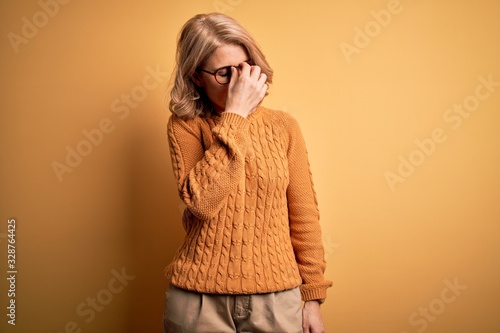 Middle age beautiful blonde woman wearing casual sweater and glasses over yellow background tired rubbing nose and eyes feeling fatigue and headache. Stress and frustration concept.