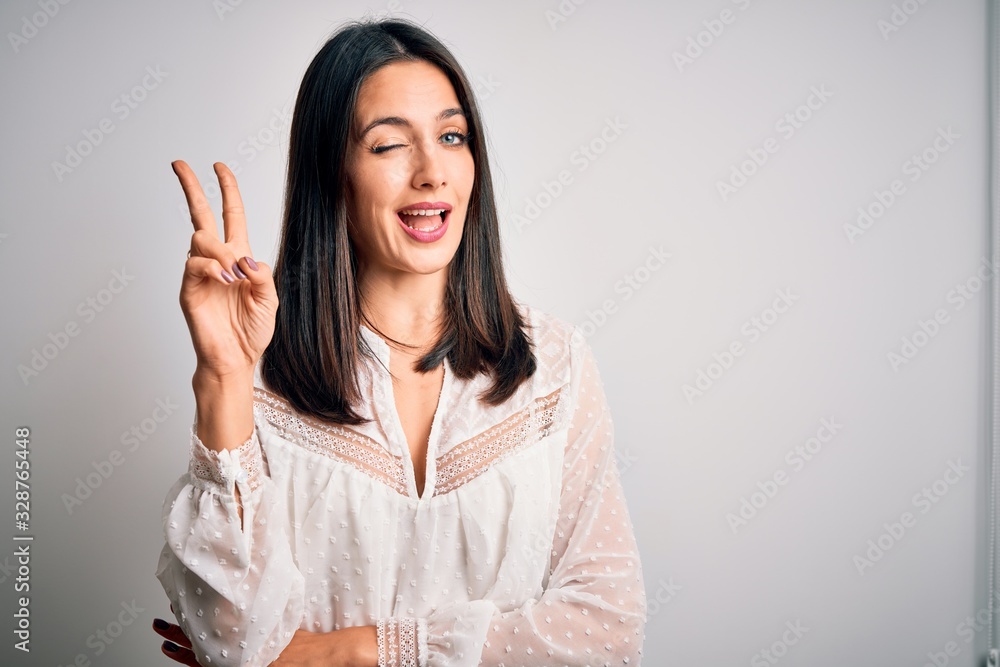 Young brunette woman with blue eyes wearing casual t-shirt over isolated white background smiling with happy face winking at the camera doing victory sign. Number two. Young brunette woman with blue eyes wearing casual t-shirt over isolated white background smiling with happy face winking at the camera doing victory sign. Number two.