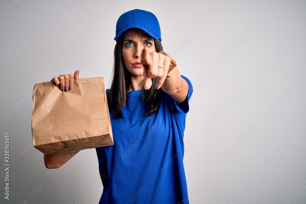 Young delivery woman with blue eyes wearing cap holding paper bag with food pointing with finger to the camera and to you, hand sign, positive and confident gesture from the front