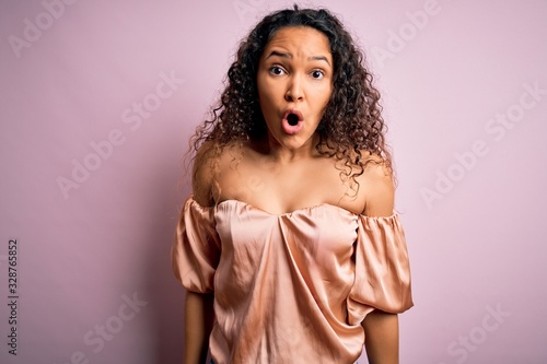Young beautiful woman with curly hair wearing casual t-shirt standing over pink background afraid and shocked with surprise and amazed expression, fear and excited face.