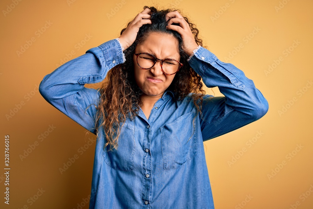Beautiful woman with curly hair wearing casual denim shirt and glasses over yellow background suffering from headache desperate and stressed because pain and migraine. Hands on head. Beautiful woman with curly hair wearing casual denim shirt and glasses over yellow background suffering from headache desperate and stressed because pain and migraine. Hands on head.