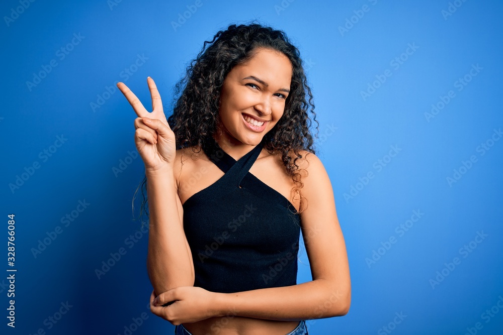 Young beautiful woman with curly hair wearing casual t-shirt standing over blue background smiling with happy face winking at the camera doing victory sign. Number two. Young beautiful woman with curly hair wearing casual t-shirt standing over blue background smiling with happy face winking at the camera doing victory sign. Number two.