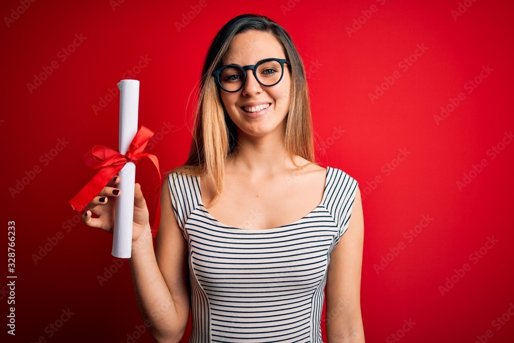 Young blonde woman with blue eyes wearing glasses holding graduated degree diploma with a happy face standing and smiling with a confident smile showing teeth Young blonde woman with blue eyes wearing glasses holding graduated degree diploma with a happy face standing and smiling with a confident smile showing teeth