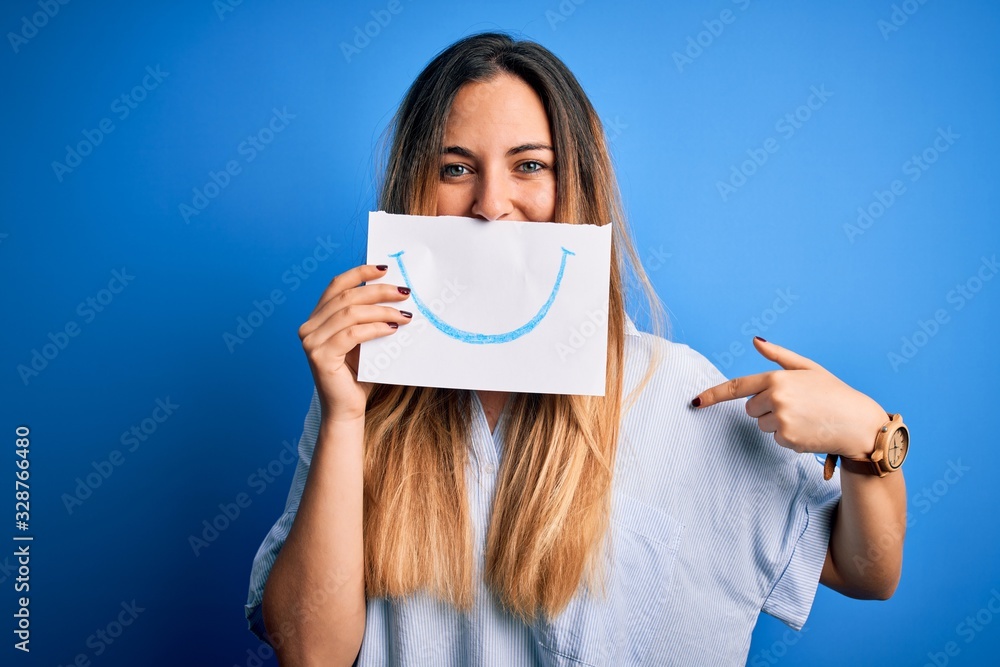 Young beautiful brunette woman holding card with smile illustration on mouth with surprise face pointing finger to himself