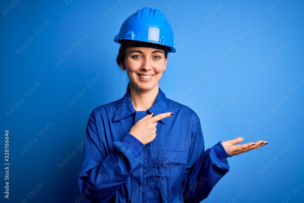 Young beautiful worker woman with blue eyes wearing security helmet and uniform amazed and smiling to the camera while presenting with hand and pointing with finger. Young beautiful worker woman with blue eyes wearing security helmet and uniform amazed and smiling to the camera while presenting with hand and pointing with finger.