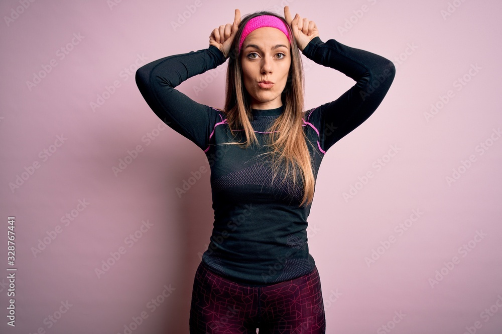 Young blonde fitness woman wearing sport workout clothes over isolated background doing funny gesture with finger over head as bull horns Young blonde fitness woman wearing sport workout clothes over isolated background doing funny gesture with finger over head as bull horns