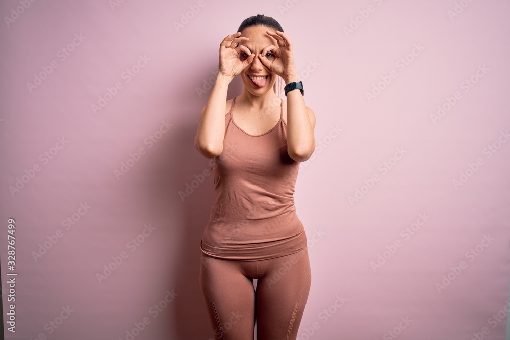 Young blonde fitness woman wearing sport workout clothes over pink isolated background doing ok gesture like binoculars sticking tongue out, eyes looking through fingers. Crazy expression. Young blonde fitness woman wearing sport workout clothes over pink isolated background doing ok gesture like binoculars sticking tongue out, eyes looking through fingers. Crazy expression.