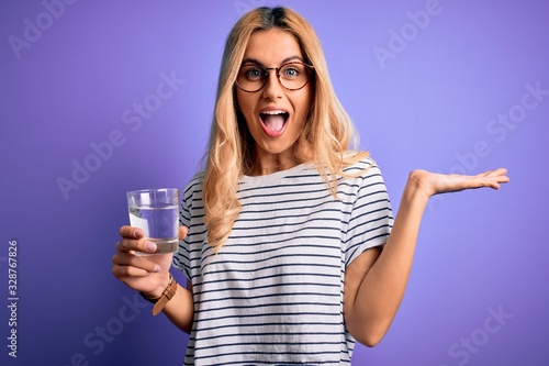 Photo Young blonde healthy woman wearing glasses drinking glass of water over purple b
