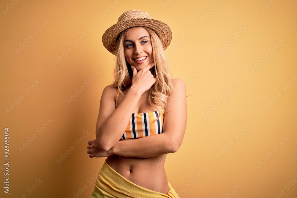 Young beautiful blonde woman on vacation wearing bikini and hat over yellow background looking confident at the camera smiling with crossed arms and hand raised on chin. Thinking positive. Young beautiful blonde woman on vacation wearing bikini and hat over yellow background looking confident at the camera smiling with crossed arms and hand raised on chin. Thinking positive.