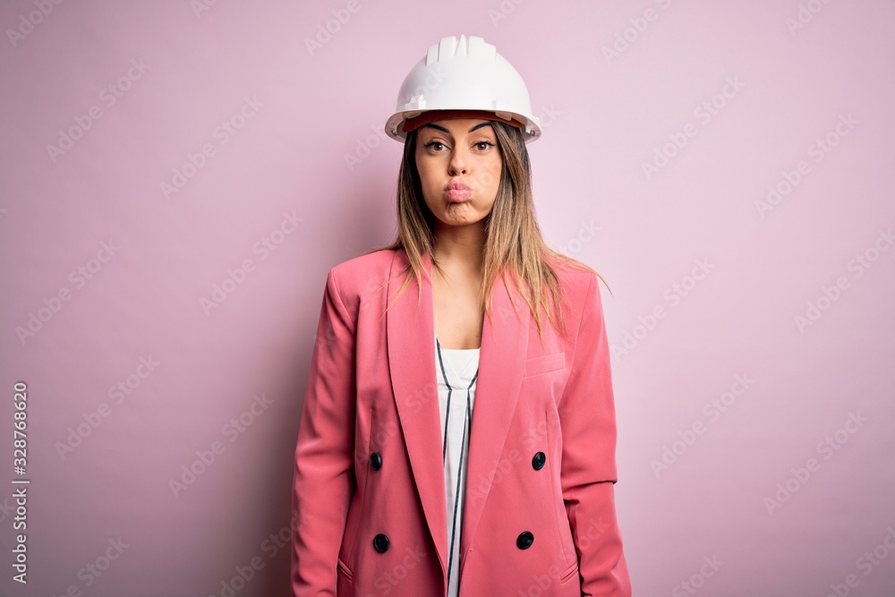Young beautiful brunette architect woman wearing safety helmet over pink background puffing cheeks with funny face. Mouth inflated with air, crazy expression. Young beautiful brunette architect woman wearing safety helmet over pink background puffing cheeks with funny face. Mouth inflated with air, crazy expression.