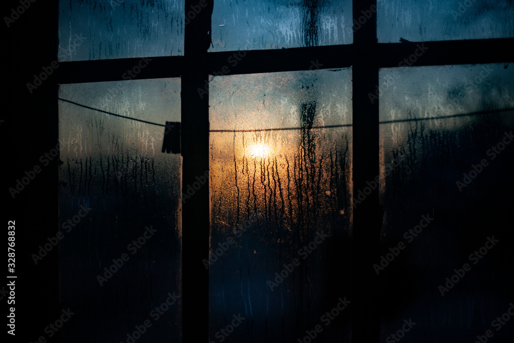 Naklejka premium Sunrise outside the window. Misted glass against the backdrop of the rising sun. Winter morning. . Simple background. The ratio of cold and warm colors. Texture of water condensate on glass.