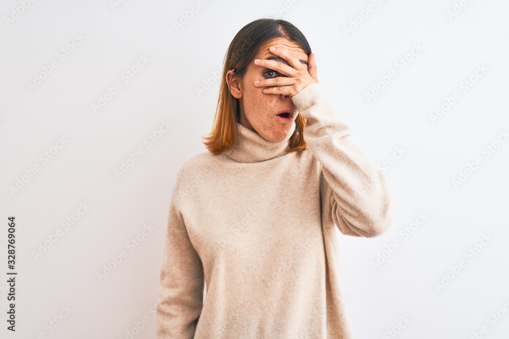 Beautiful redhead woman wearing winter turtleneck sweater over isolated background peeking in shock covering face and eyes with hand, looking through fingers with embarrassed expression. Beautiful redhead woman wearing winter turtleneck sweater over isolated background peeking in shock covering face and eyes with hand, looking through fingers with embarrassed expression.