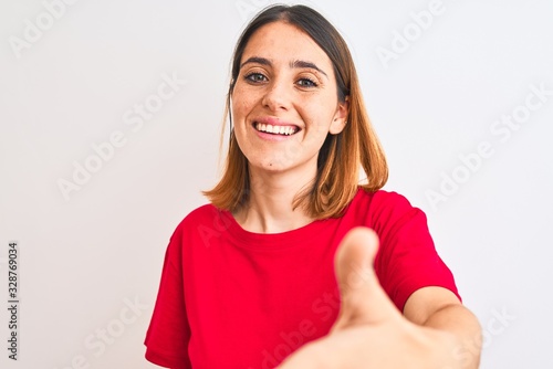 Beautiful redhead woman wearing casual red t-shirt over isolated background smiling friendly offering handshake as greeting and welcoming. Successful business.