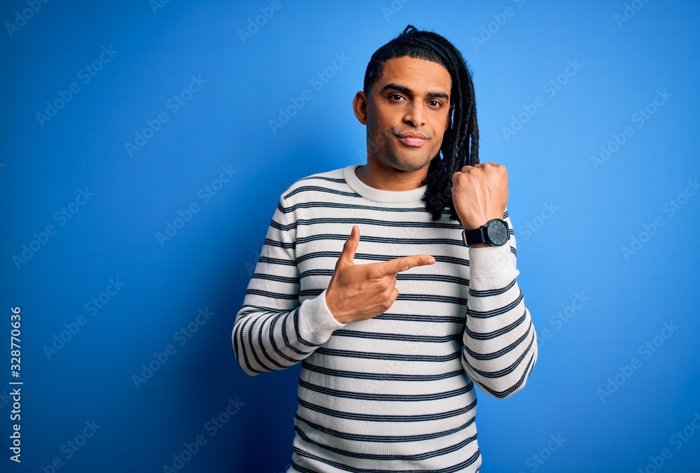 Fototapeta premium Young handsome african american afro man with dreadlocks wearing casual striped sweater In hurry pointing to watch time, impatience, looking at the camera with relaxed expression