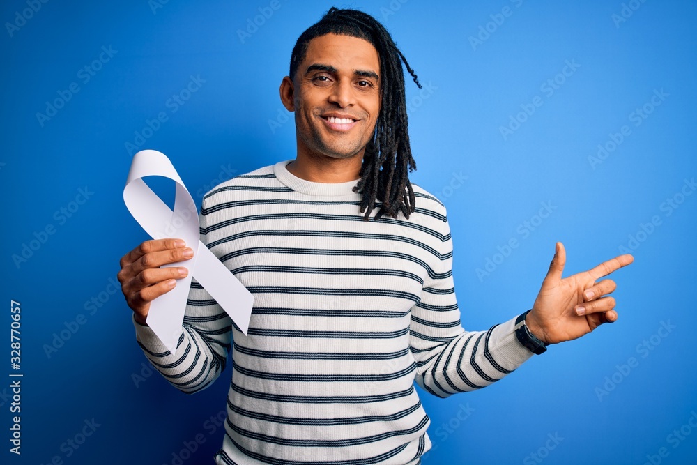 Young african american man with dreadlocks holding white ribbon over blue background very happy pointing with hand and finger to the side Young african american man with dreadlocks holding white ribbon over blue background very happy pointing with hand and finger to the side