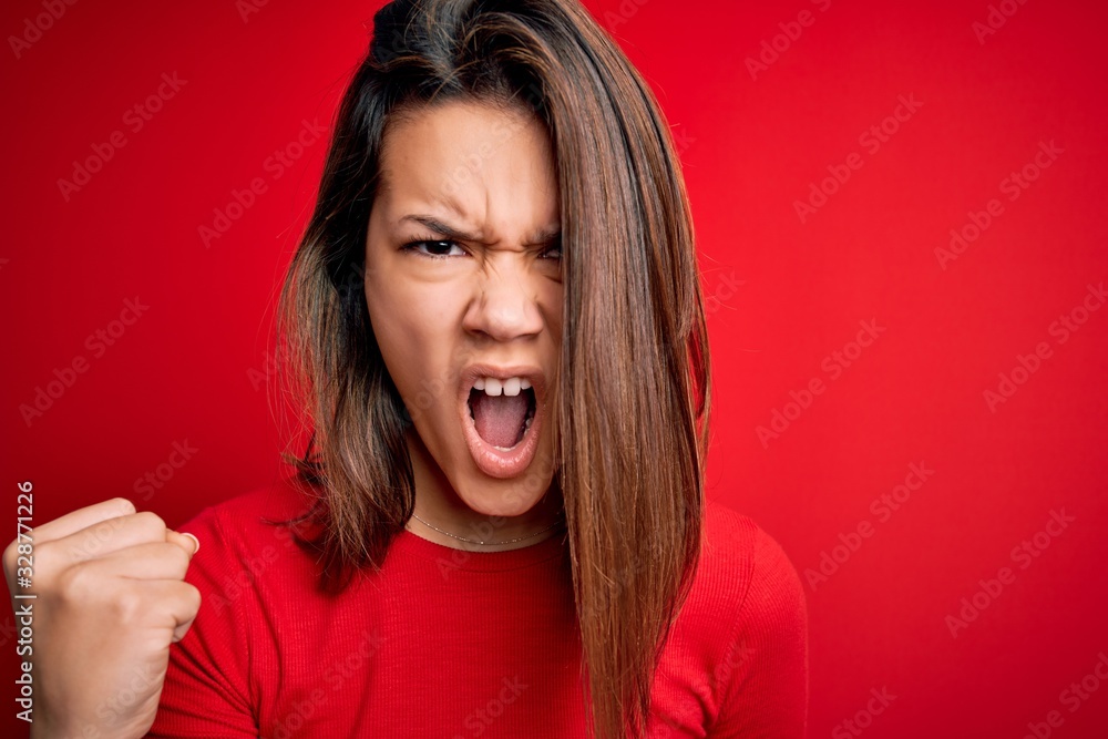 Young beautiful brunette girl wearing casual t-shirt over isolated red background angry and mad raising fist frustrated and furious while shouting with anger. Rage and aggressive concept. Young beautiful brunette girl wearing casual t-shirt over isolated red background angry and mad raising fist frustrated and furious while shouting with anger. Rage and aggressive concept.