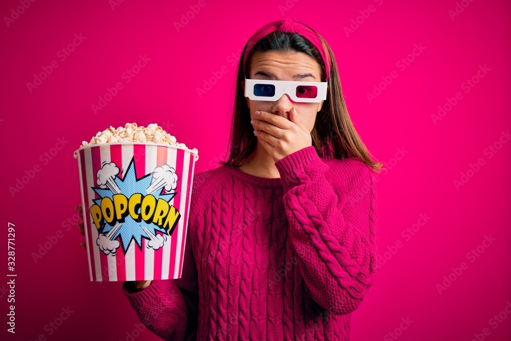 Obraz premium Young beautiful girl watching movie using 3d glasses eating box with popcorns shocked covering mouth with hands for mistake. Secret concept.