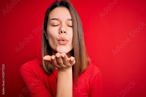 Young beautiful brunette girl wearing casual t-shirt over isolated red background looking at the camera blowing a kiss with hand on air being lovely and sexy. Love expression. © Krakenimages.com