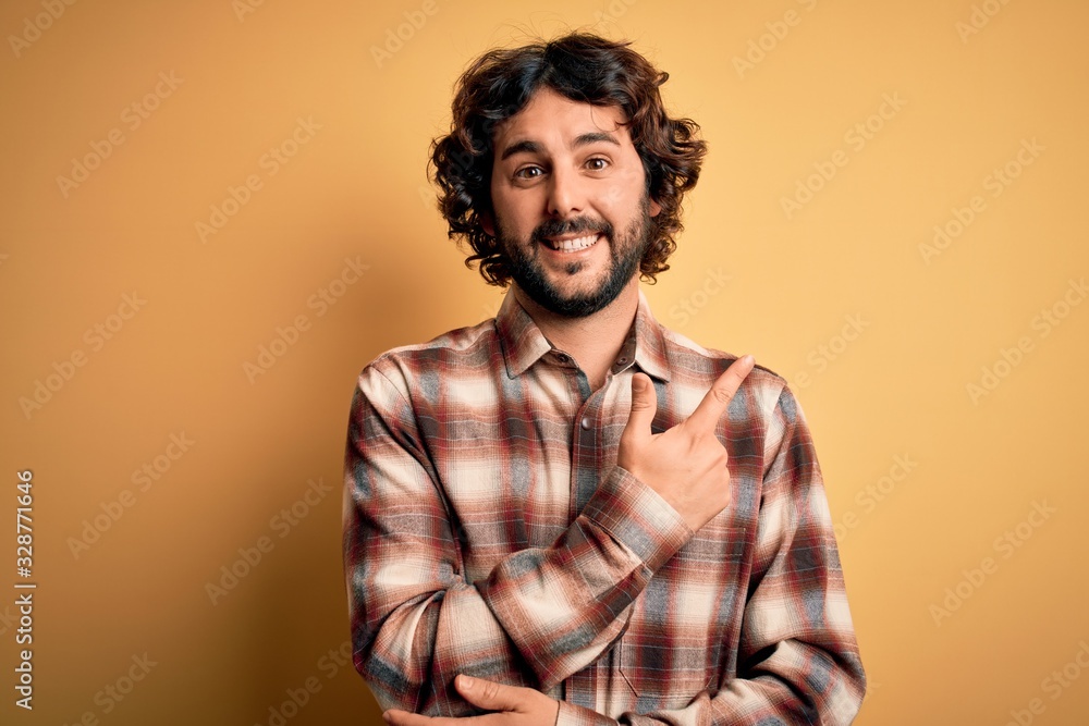 Young handsome man with beard wearing casual shirt standing over yellow background cheerful with a smile of face pointing with hand and finger up to the side with happy and natural expression on face Young handsome man with beard wearing casual shirt standing over yellow background cheerful with a smile of face pointing with hand and finger up to the side with happy and natural expression on face