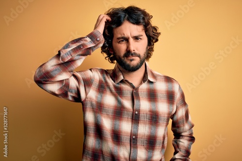 Young handsome man with beard wearing casual shirt standing over yellow background confuse and wonder about question. Uncertain with doubt, thinking with hand on head. Pensive concept.