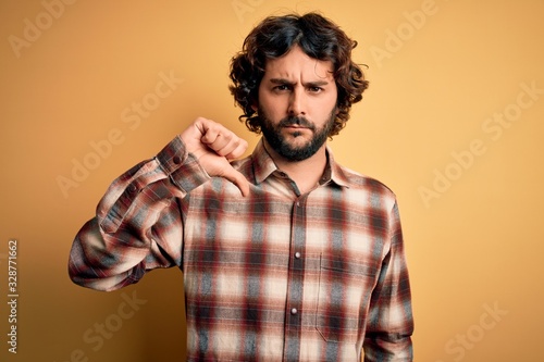 Young handsome man with beard wearing casual shirt standing over yellow background looking unhappy and angry showing rejection and negative with thumbs down gesture. Bad expression.