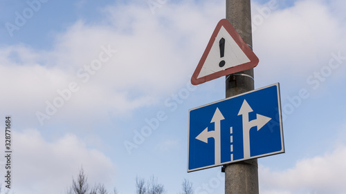 Road sign on a pillar against a blue sky with clouds. Signs of an emergency hazardous area and lane directions.