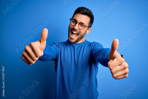 Young handsome man with beard wearing casual sweater and glasses over blue background approving doing positive gesture with hand, thumbs up smiling and happy for success. Winner gesture. photo