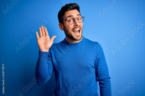 Young handsome man with beard wearing casual sweater and glasses over blue background Waiving saying hello happy and smiling, friendly welcome gesture photo