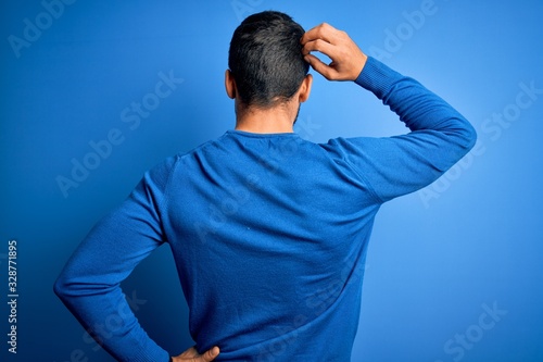 Young handsome man with beard wearing casual sweater and glasses over blue background Backwards thinking about doubt with hand on head