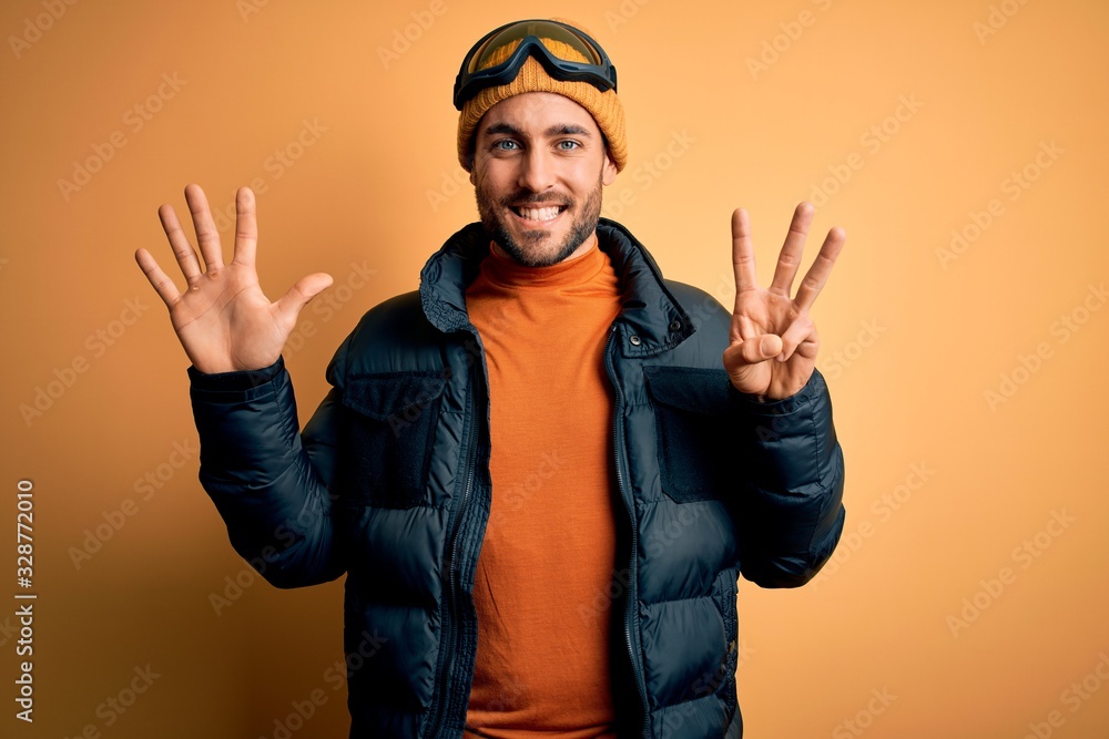 Young handsome skier man with beard wearing snow sportswear and ski goggles showing and pointing up with fingers number eight while smiling confident and happy. Young handsome skier man with beard wearing snow sportswear and ski goggles showing and pointing up with fingers number eight while smiling confident and happy.