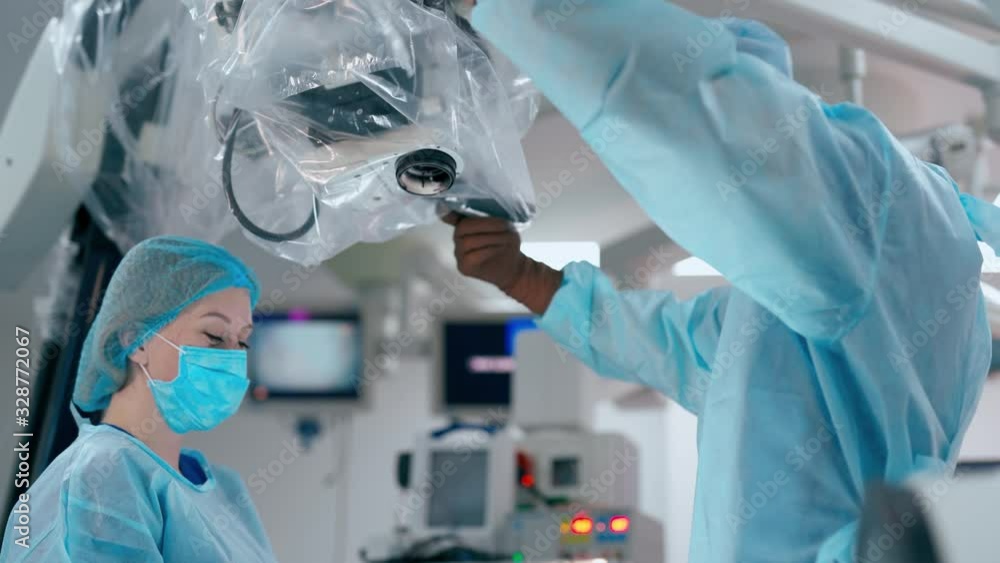 Doctor and his assistant in the operating room. Specialist in a medical uniform customizes microscope before the operation. Medical concept. Doctor and his assistant in the operating room. Specialist in a medical uniform customizes microscope before the operation. Medical concept.