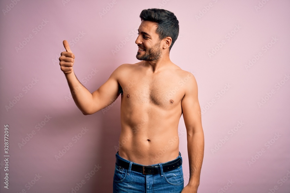 Young handsome strong man with beard shirtless standing over isolated pink background Looking proud, smiling doing thumbs up gesture to the side