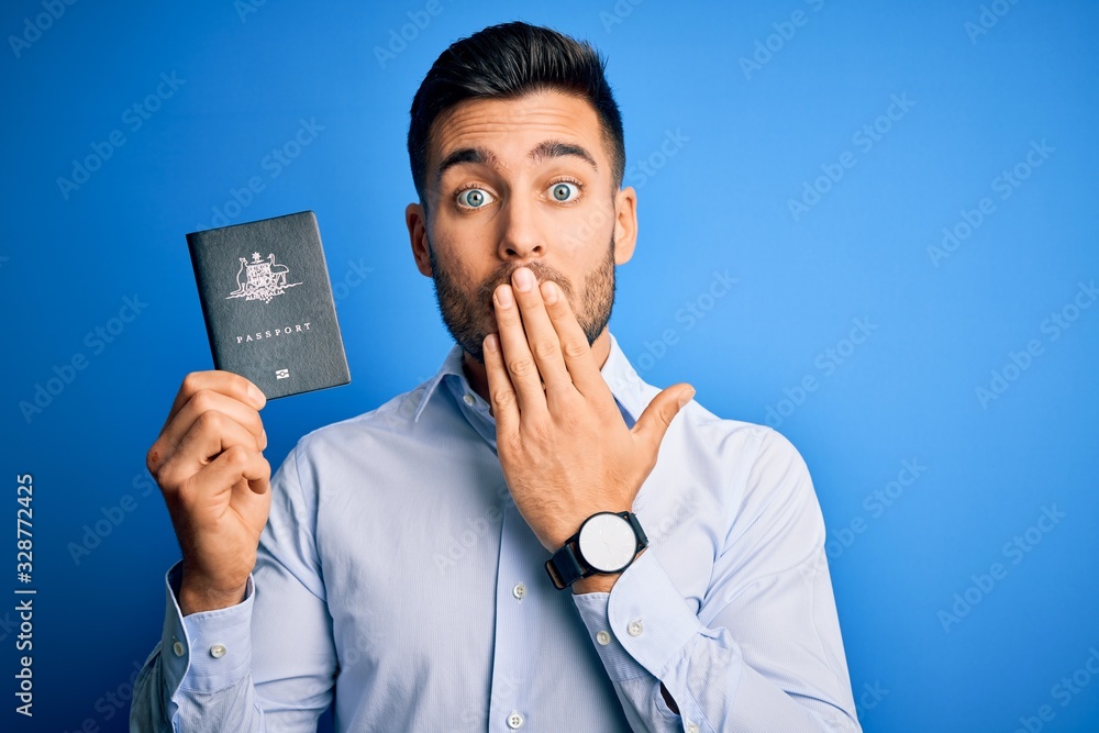 Young handsome tourist man holding australia australian passport id over blue background cover mouth with hand shocked with shame for mistake, expression of fear, scared in silence, secret concept Young handsome tourist man holding australia australian passport id over blue background cover mouth with hand shocked with shame for mistake, expression of fear, scared in silence, secret concept