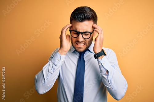 Young handsome businessman wearing tie and glasses standing over yellow background with hand on headache because stress. Suffering migraine.