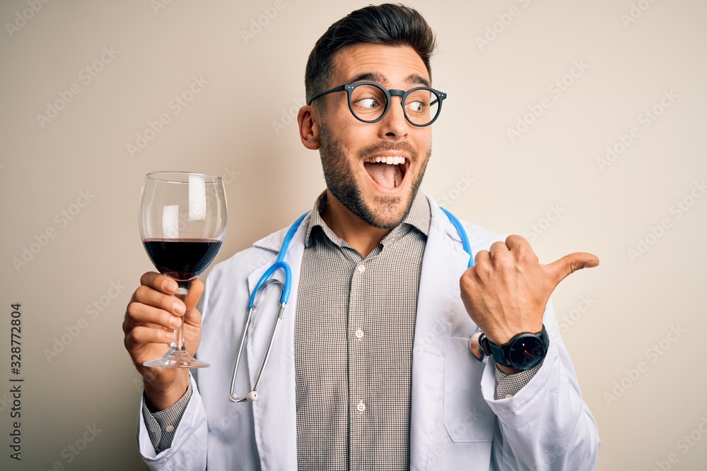 Young doctor man wearing stethoscope drinking a glass of fresh wine over isolated background pointing and showing with thumb up to the side with happy face smiling Young doctor man wearing stethoscope drinking a glass of fresh wine over isolated background pointing and showing with thumb up to the side with happy face smiling