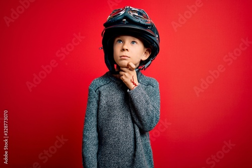 Young little caucasian kid wearing vintage biker motorcycle helmet and googles over red background with hand on chin thinking about question, pensive expression. Smiling with thoughtful face. Doubt
