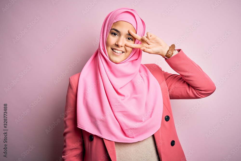 Young beautiful girl wearing muslim hijab standing over isolated pink background Doing peace symbol with fingers over face, smiling cheerful showing victory Young beautiful girl wearing muslim hijab standing over isolated pink background Doing peace symbol with fingers over face, smiling cheerful showing victory