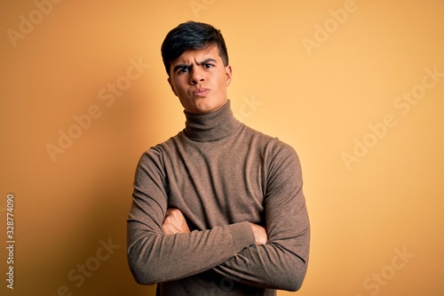 Young handsome man wearing casual turtleneck sweater over isolated yellow background skeptic and nervous, disapproving expression on face with crossed arms. Negative person. © Krakenimages.com