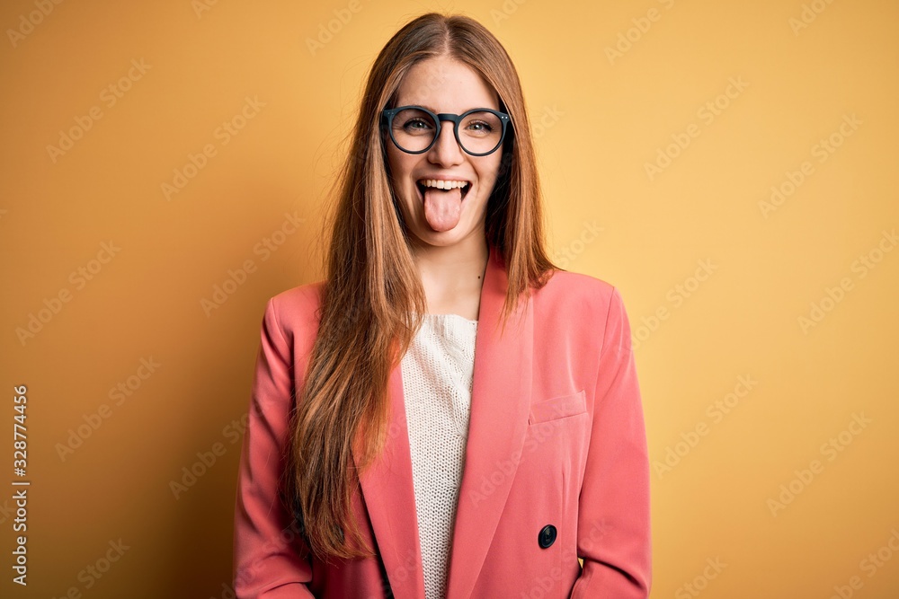 Young beautiful redhead woman wearing jacket and glasses over isolated yellow background sticking tongue out happy with funny expression. Emotion concept.