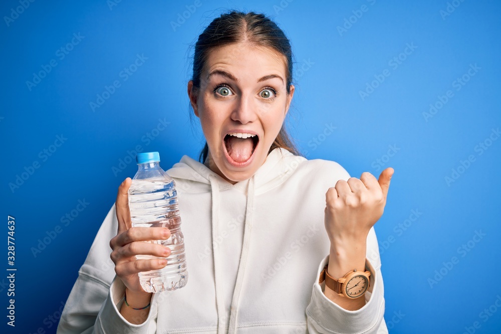 Young beautiful redhead woman doing sport drinking bottle of water over blue background screaming proud and celebrating victory and success very excited, cheering emotion Young beautiful redhead woman doing sport drinking bottle of water over blue background screaming proud and celebrating victory and success very excited, cheering emotion