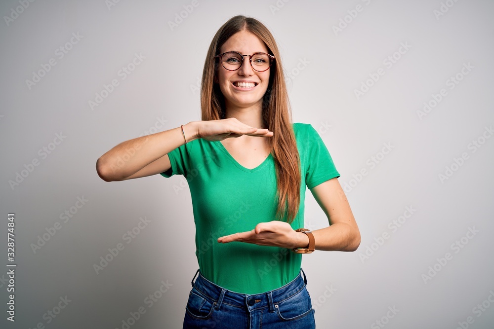 Young beautiful redhead woman wearing casual green t-shirt and glasses over white background gesturing with hands showing big and large size sign, measure symbol. Smiling looking at the camera Young beautiful redhead woman wearing casual green t-shirt and glasses over white background gesturing with hands showing big and large size sign, measure symbol. Smiling looking at the camera