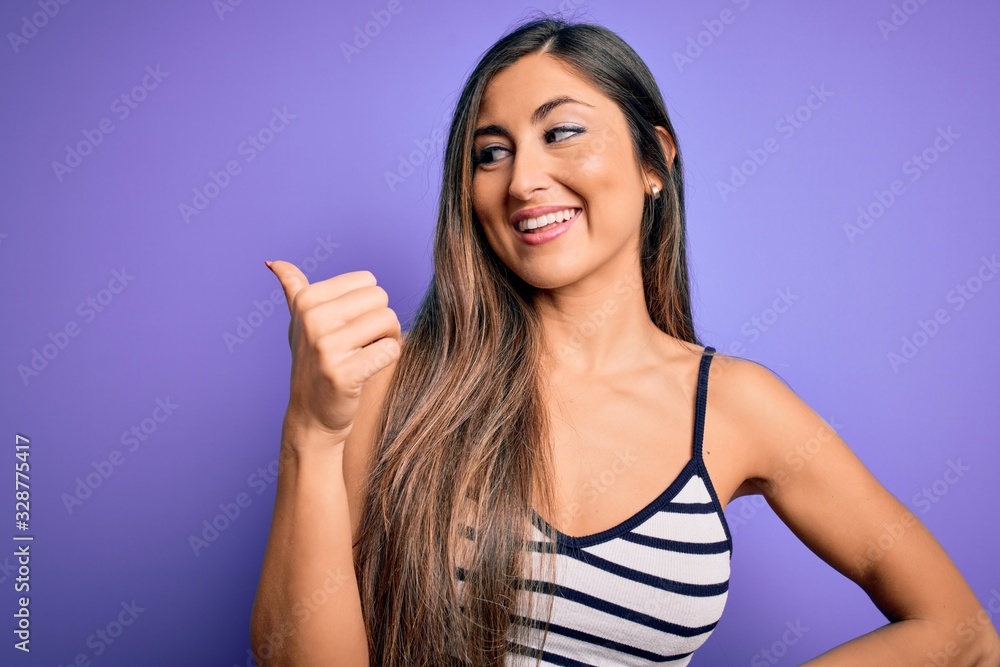 Young beautiful woman wearing casual summer striped top over purple isolated background smiling with happy face looking and pointing to the side with thumb up. Young beautiful woman wearing casual summer striped top over purple isolated background smiling with happy face looking and pointing to the side with thumb up.
