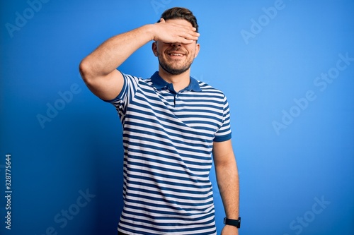 Young man with blue eyes wearing glasses and casual striped t-shirt over blue background smiling and laughing with hand on face covering eyes for surprise. Blind concept.