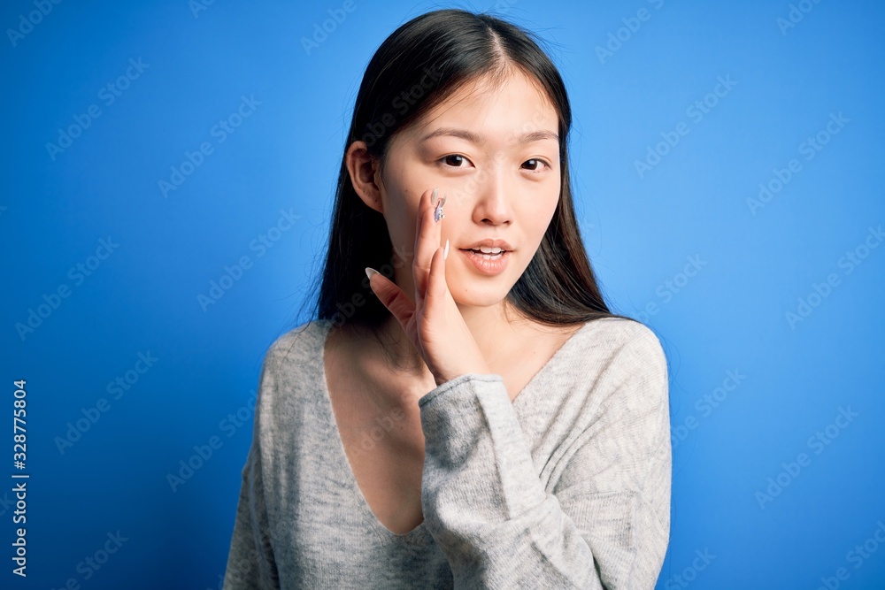 Young beautiful asian woman wearing casual sweater standing over blue isolated background hand on mouth telling secret rumor, whispering malicious talk conversation Young beautiful asian woman wearing casual sweater standing over blue isolated background hand on mouth telling secret rumor, whispering malicious talk conversation