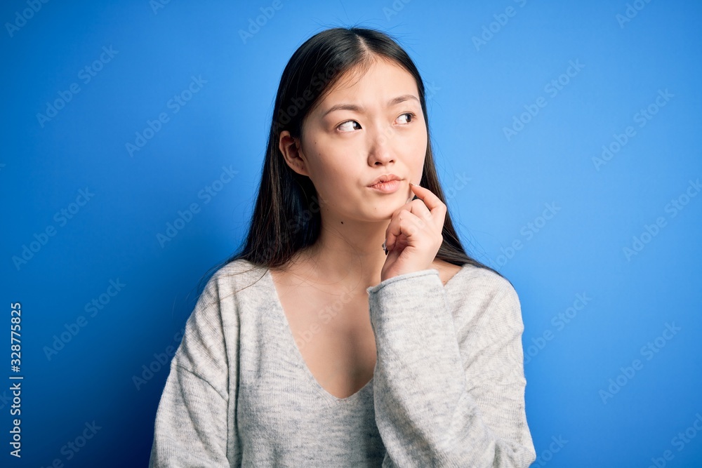 Young beautiful asian woman wearing casual sweater standing over blue isolated background with hand on chin thinking about question, pensive expression. Smiling with thoughtful face. Doubt concept. Young beautiful asian woman wearing casual sweater standing over blue isolated background with hand on chin thinking about question, pensive expression. Smiling with thoughtful face. Doubt concept.